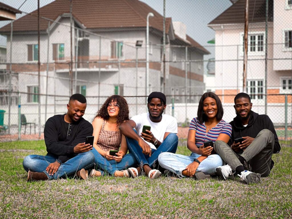 Five young adults sit on grass in front of a chain-link fence and houses, smiling and holding their phones, enjoying time together outdoors.
